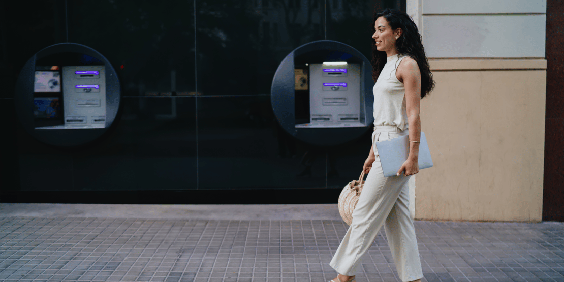 Smiling woman carrying a laptop and handbag while walking past outdoor ATMs