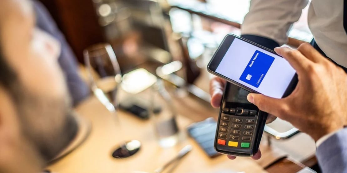 Person using a smartphone with a digital card displayed to make a contactless payment on a handheld card terminal in a restaurant setting.